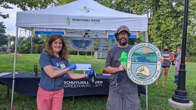 A woman and a man stand in front a tent holding a Schuylkill River Greenways sign.