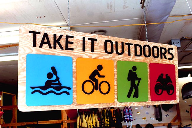 A large plywood sign that says Take it outdoors with stick figure people on bikes and kayaks against a blue, red, green and yellow backcground.