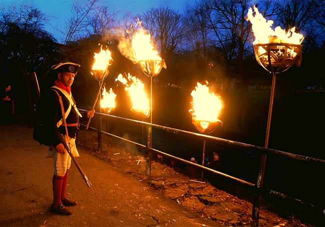 A man in Continental Army soldier uniform holds a lighted stick near other lit fires.