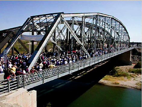 A mass of people stand on a metal bridge over a green river.