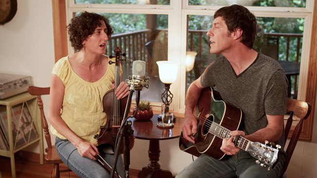 A woman in yellow with curly brown hair. holding a violin, sings with a man in grey holding a guitar.
