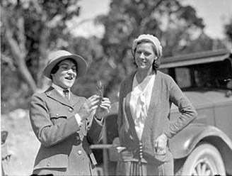 Pauline Mead in her NPS uniform and a soft-brimmed hat shows a visitor a plant.