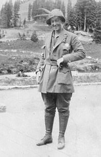 Smiling Alma Wagen stands in her company guide outfit, guide badge, and hat with two long feathers.