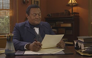 A black man wearing a blue suit sits at a desk holding a piece of paper
