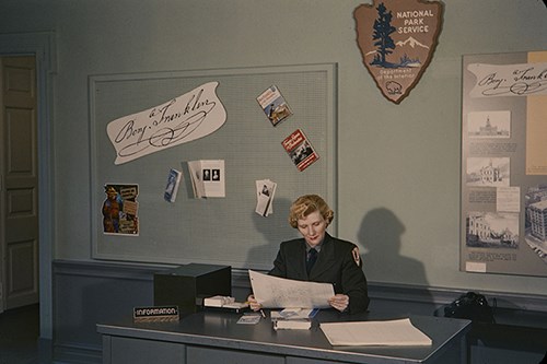 Unidentified woman wearing an NPS uniform sits at an information desk, reading a page from the park's visitor log.