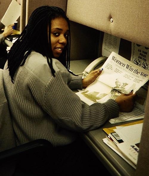 Unidentified woman in the NPS cardigan sitting at a desk reading a newspaper.