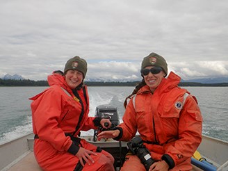 Two smiling women in orange jumpsuits and green knit caps travel by boat to conduct fieldwork.