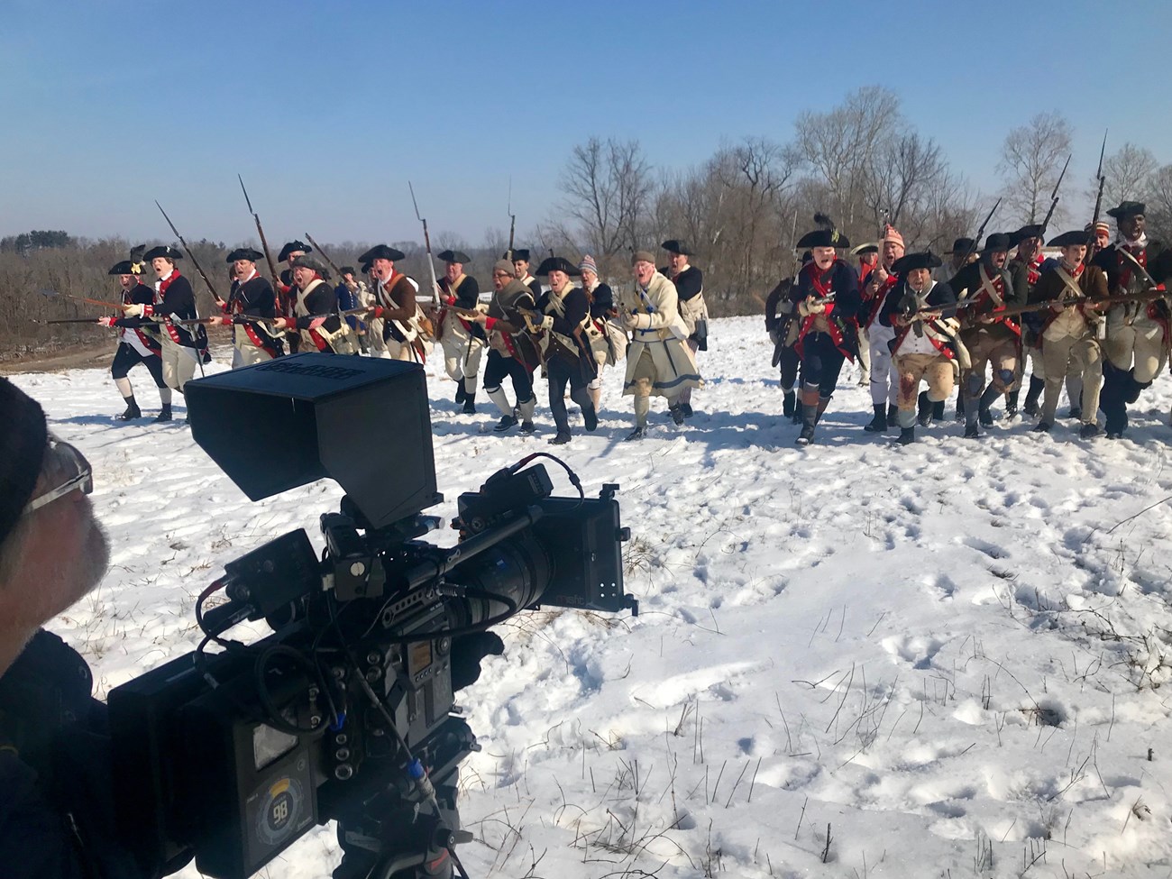 A man holds a camera towards a group of people dressed in revolutionary war costumes coming towards us.