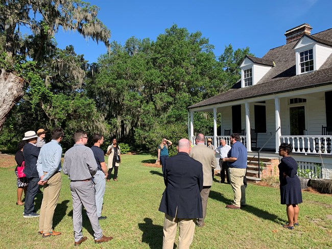 A mixed-race group of professionals stands in a circle before an old plantation house and a live oak tree.