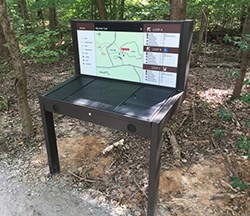 A brown sign with a tactile map and brown and white directional trail signs.