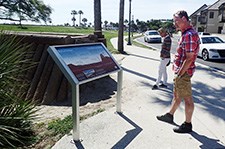 A man in shorts stands on the sidewalk with his hands in his pockets as he reads a low profile wayside.