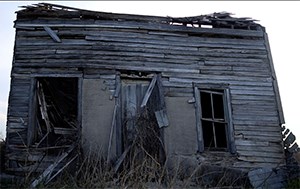 A dilapidated  wooden house stands with the roof falling in and the the left side window missing