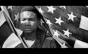 A young black man holds an American flag with the stars framed behind his head as we he looks forward in solemn determination