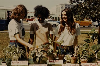 Three women at a Summer in the Parks plant sale. The middle women wears the 1970 beige uniform.