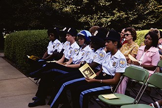 Six women in US Park Police uniform sit in folding chairs and hold paper programs in their hands.