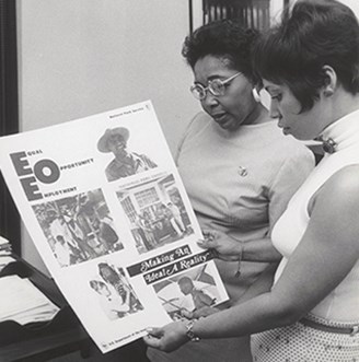 Two black women stand to the right of the image looking at an EEO poster. One woman is wearing a light colored sleeveless top with a short haircut. The other woman wears a longer sleeved top and a pin on her left chest.