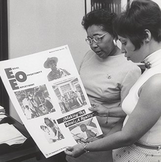 Two black women stand to the right of the image looking at an EEO poster. One woman is wearing a light colored sleeveless top with a short haircut. The other woman wears a longer sleeved top and a pin on her left chest. Two black women stand to the right of the image looking at an EEO poster. One woman is wearing a light colored sleeveless top with a short haircut. The other woman wears a longer sleeved top and a pin on her left chest.