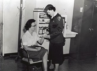 Olive Johnson in her NPS uniform stands over a seated woman, bandaging wound on her hand.