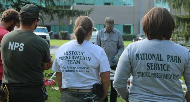 lt= NPS employees standing around an instructor. The back of their t-shirts read "Historic Preservation Team."