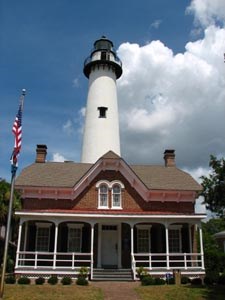 St. Simon's Lighthouse on a sunny day, with blue sky and fluffy white clouds. The white tower rises behind the 2-story red brick keeper's house, which has a white porch.