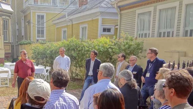 A group of people gathered around a speaker in a courtyard surrounded by historic wood-frame houses.