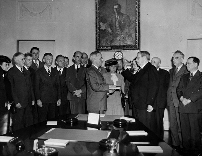 Truman's Inauguration, April 12, 1945 A crowd gathered at the end of a long table in a room. A man has his hand on a book during the presidential swearing-in ceremony