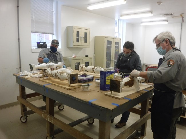 Three people wearing face masks and gloves stand around a work bench repairing wooden balusters. Three people wearing face masks and gloves stand around a work bench repairing wooden balusters.