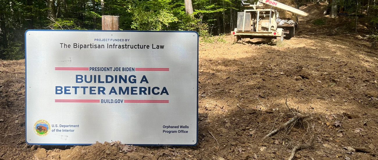 Building a Better America a white sign sits in a tree lined dirt lot, the sign reads "Project Funded by The Bipartisan Infrastructure Law - Building a Better America"