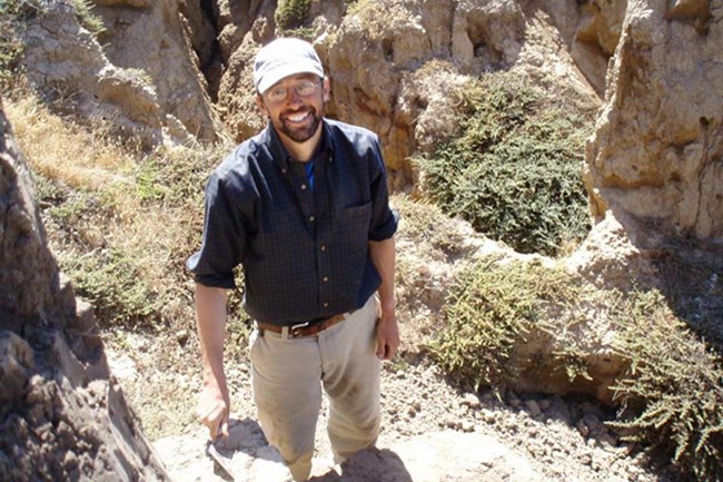 Man with blue shirt and white hat standing next to rock formation.