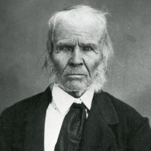 lt= Historic black and white photo of elderly man with white hair in black jacket.