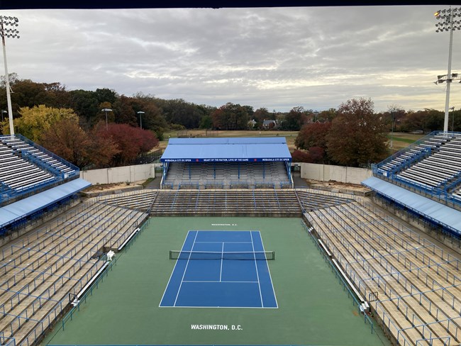 Aerial image looking down on center court at the Rock Creek Tennis Center which is surounded by bleacher seats on all sides with a clouded sky and trees off in the distance.