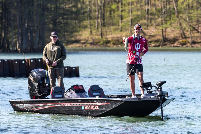 two men stand on top of a boat holding fishing poles with a forest in the background. The man on the right is smiling while holding a green and white fish.