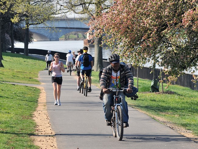 lt= Visitors ride bikes and jog down a paved path along a river in the Georgetown Waterfront Park during the springtime.