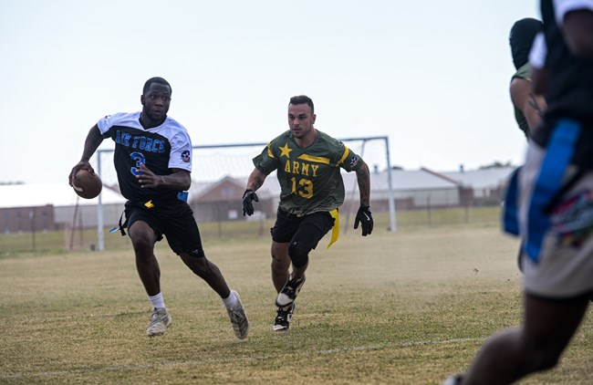 U.S. Air Force On a dusty football field two men run towards the left side of the photo. The man in front holds a football wearing a black and white shirt with the words "Air Force" on it. The man close behind wears a green and yellow shirt with the words "Army 13" on i