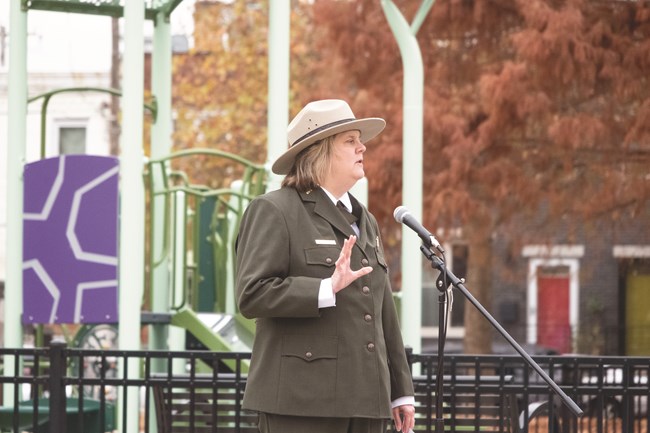 Mifflin Square - ORLP Grant Ribbon Cutting A female ranger wearing a green formal button down uniform and felt flat rimmed hat holds her hand up as she speaks into a microphone with a colorful children’s playground in the background.