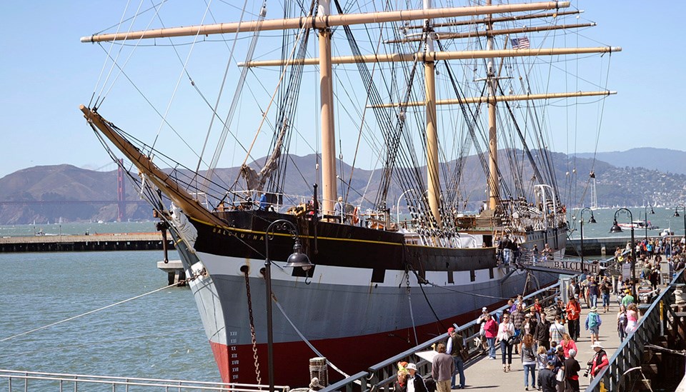 People walk by a three-masted ship docked alongside pier