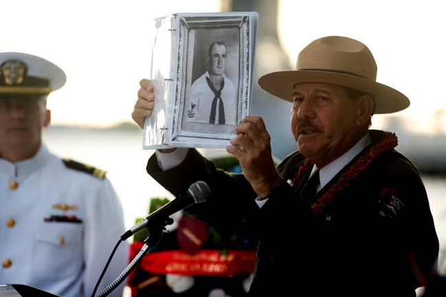 Park ranger at a podium holds up a photo of a person in a navy uniform. He is surrounded by people in navy dress uniforms.