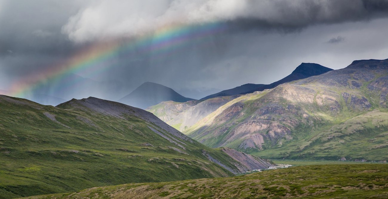 Photo of mountains with arctic tundra under cloudy skies and a rainbow.