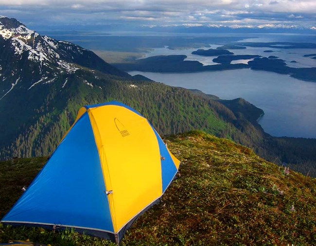 A tent is perched on a mountain peak overlooking a large lake.