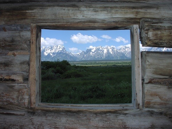 A cabin window frames a view looking into a pasture and snow capped mountains beyond