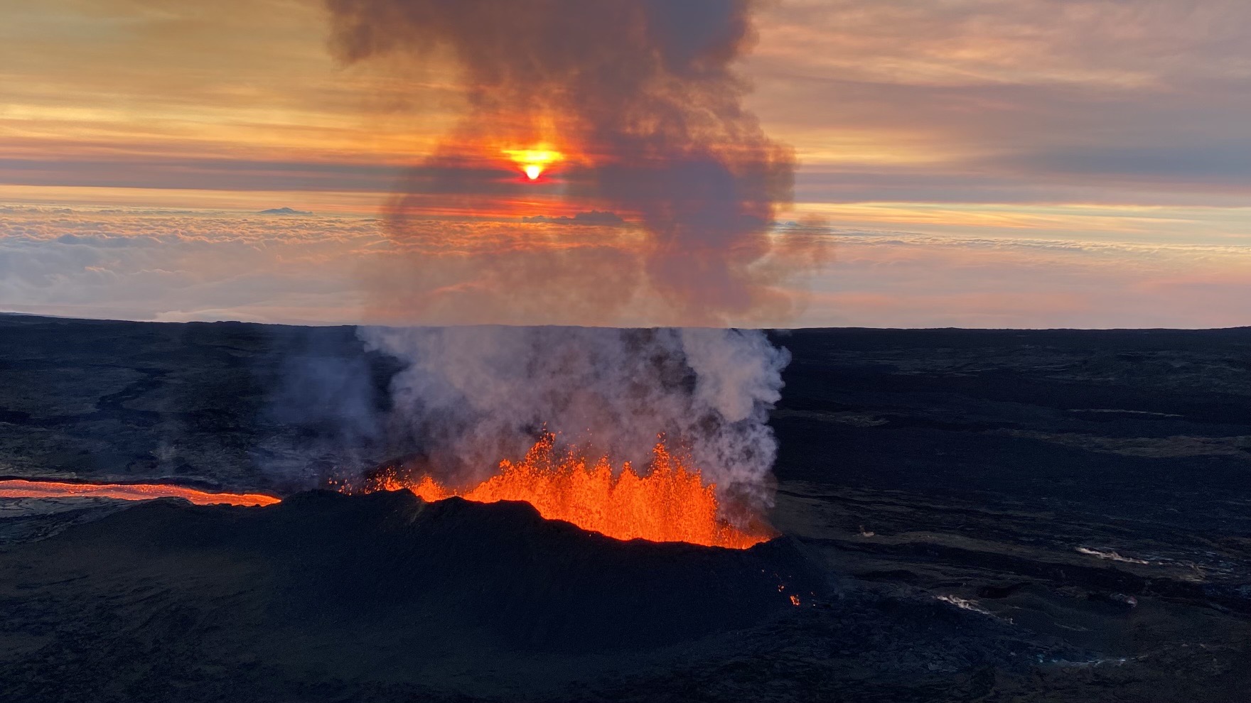 Lava flows like a river below volcano and erupts from volcano - the orange lava contrasting against the black of former flows. At sunrise.