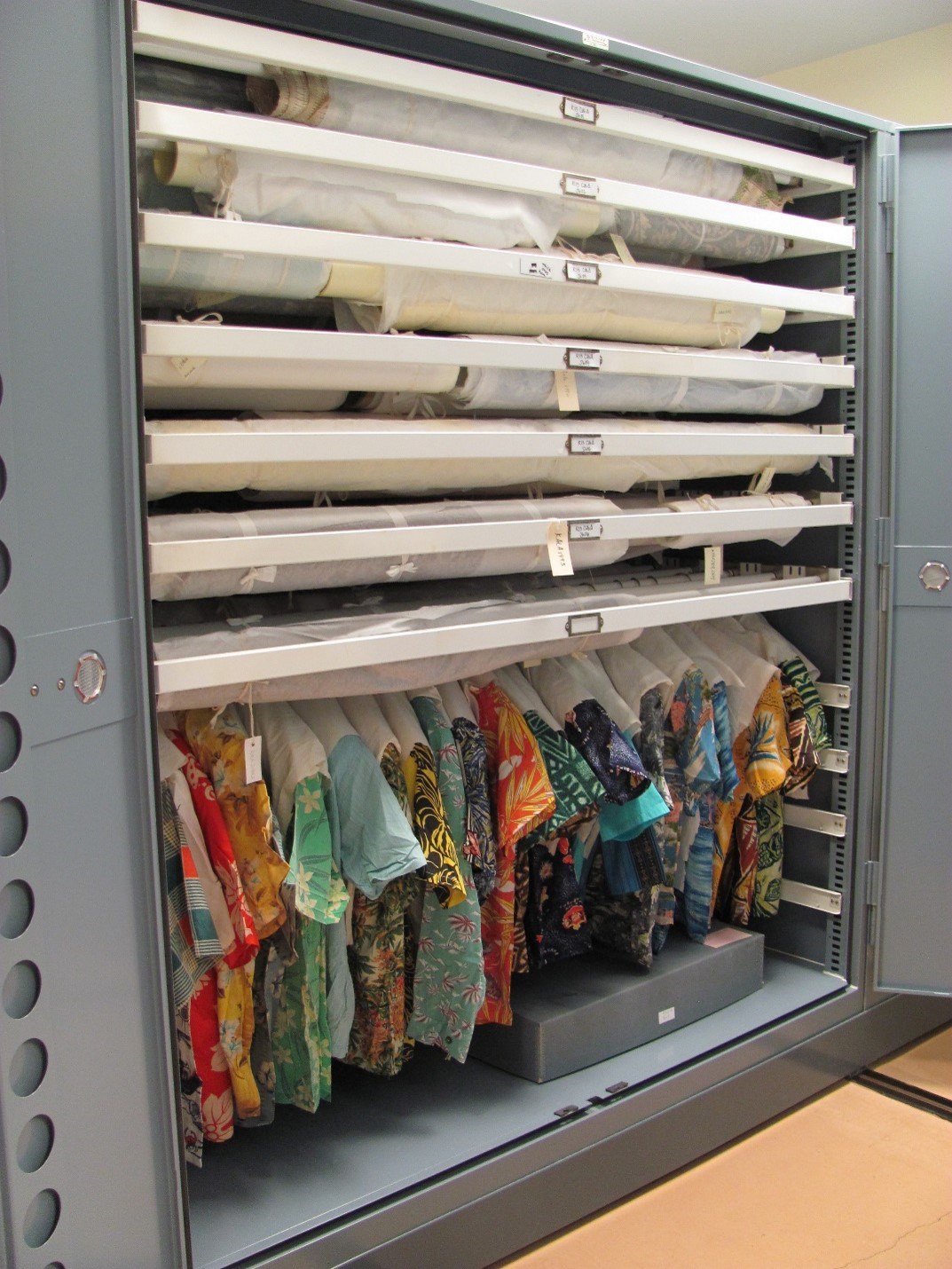 Large gray cabinet with colorful shirts handing in the bottom and rolled textiles above.