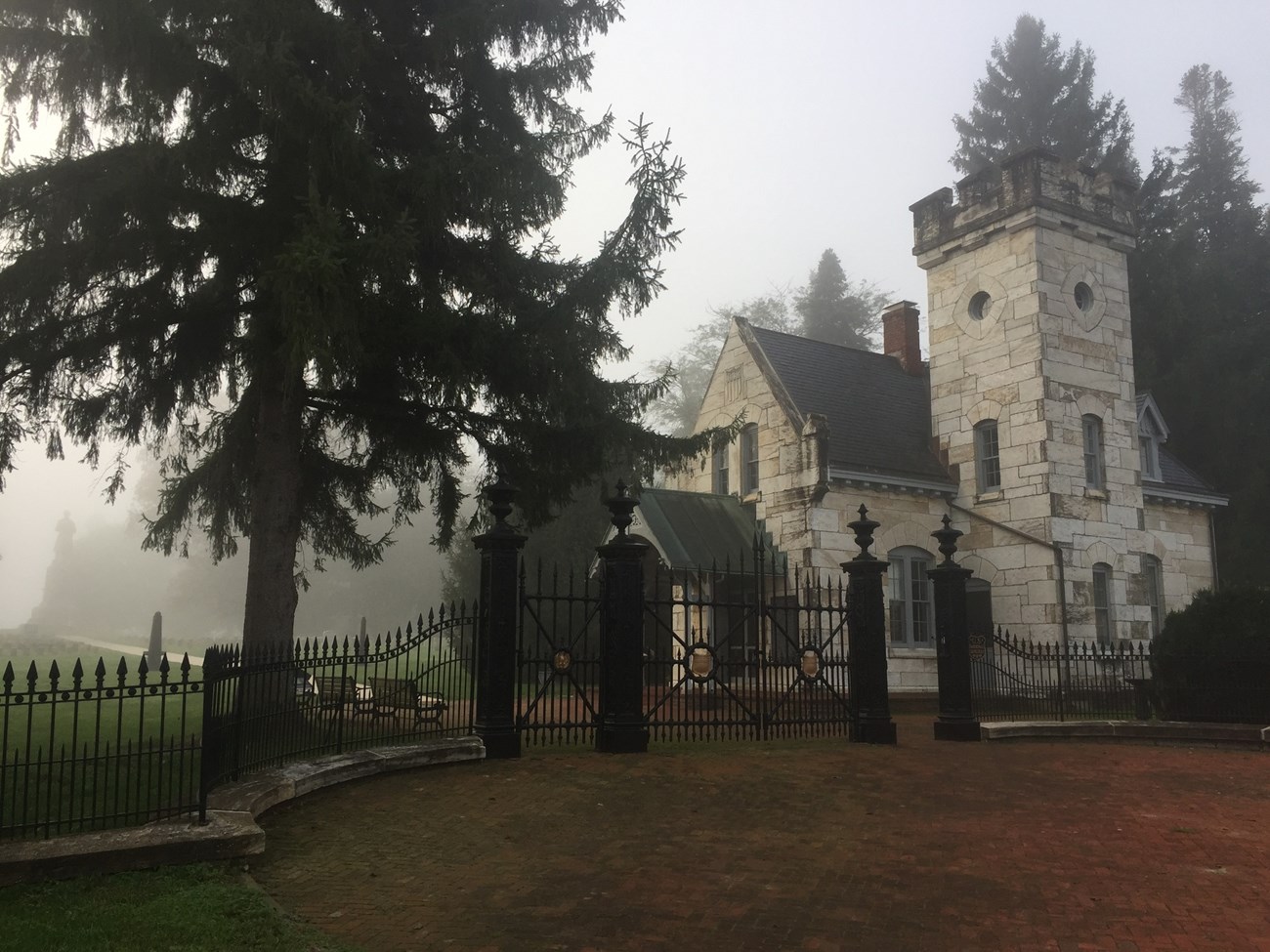 Lodge and gate and Antietam National Cemetery A gothic stone lodge stands just inside the gate of an ornate black iron fence, with a foggy cemetery behind
