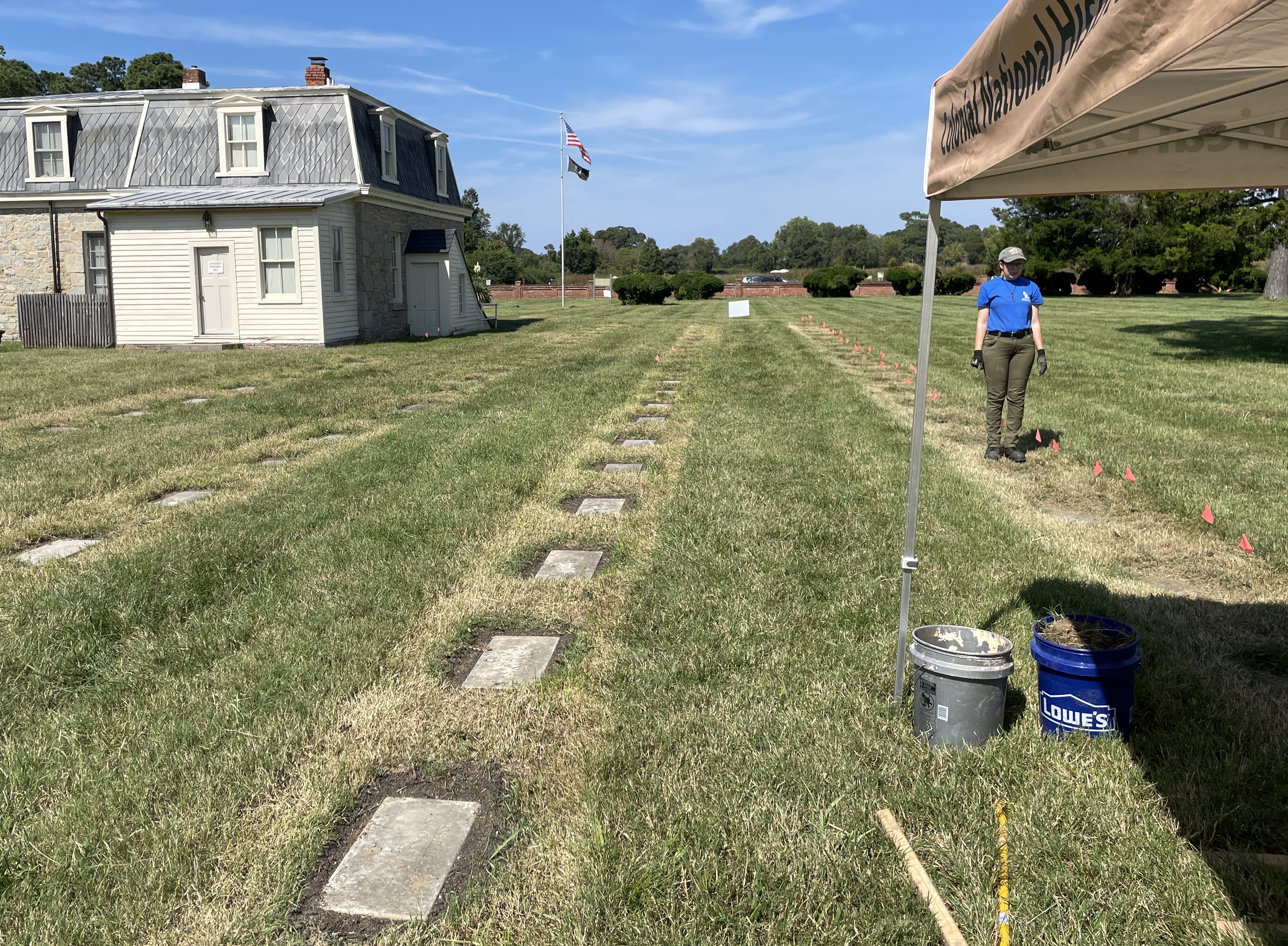 A crew member stands near a row of flags marking headstone locations, to the right of a completed row of leveled and lifted markers
