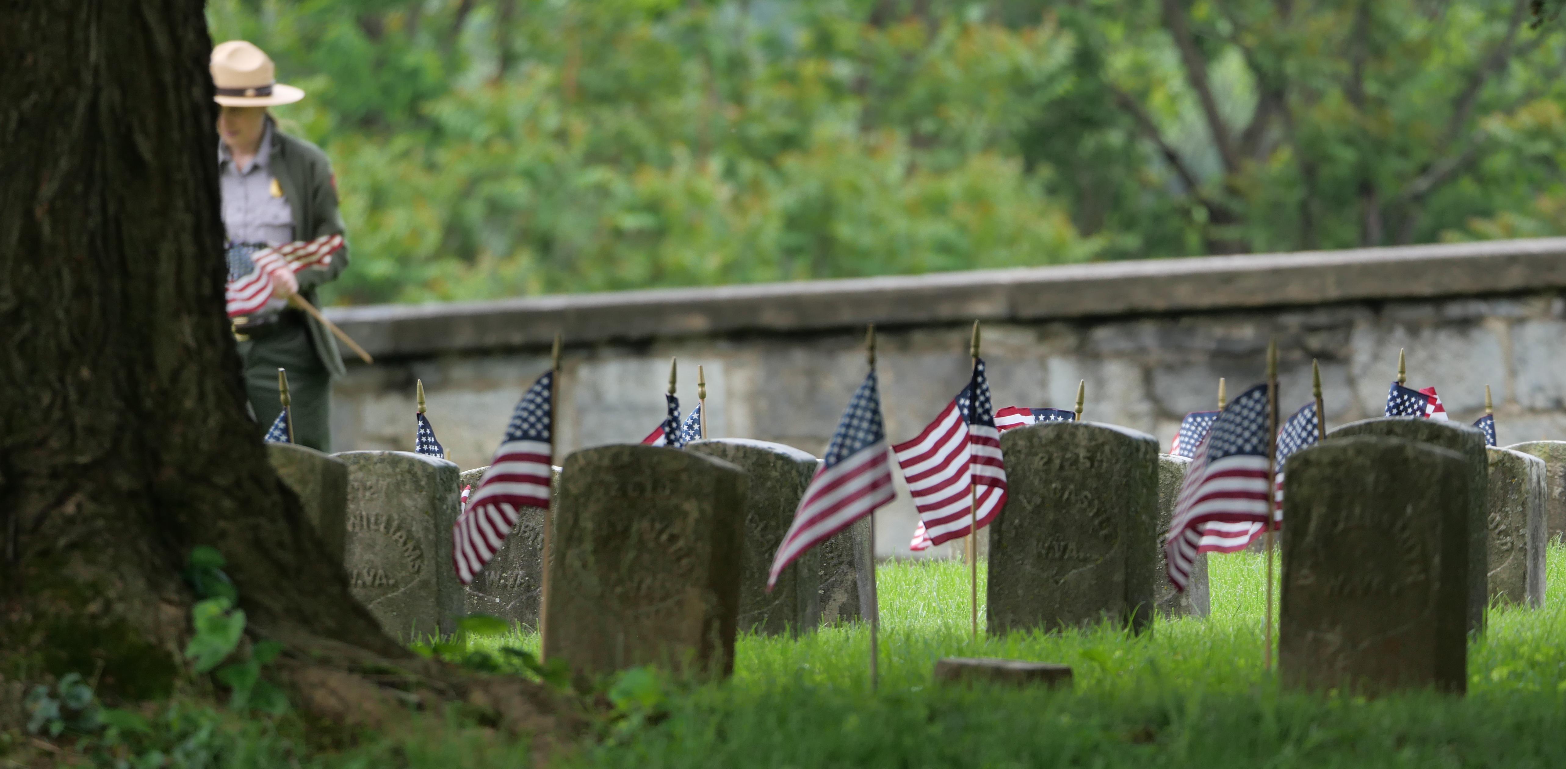 American flags are placed beside grave markers near a tall tree and stone wall.