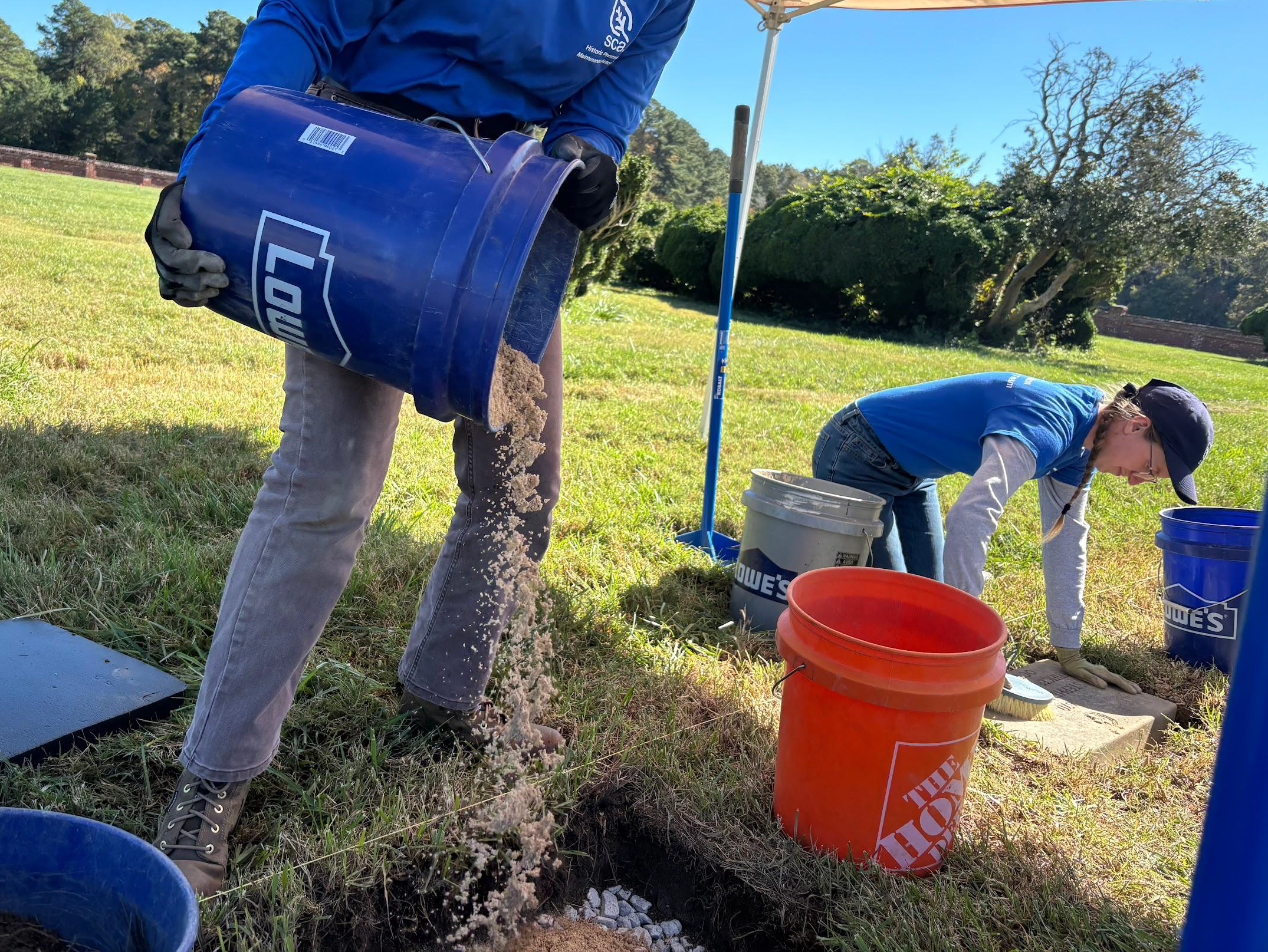 One crew member pours sand from a 5-gallon bucket into a hole, as another person kneels with a soft brush to clean a marker