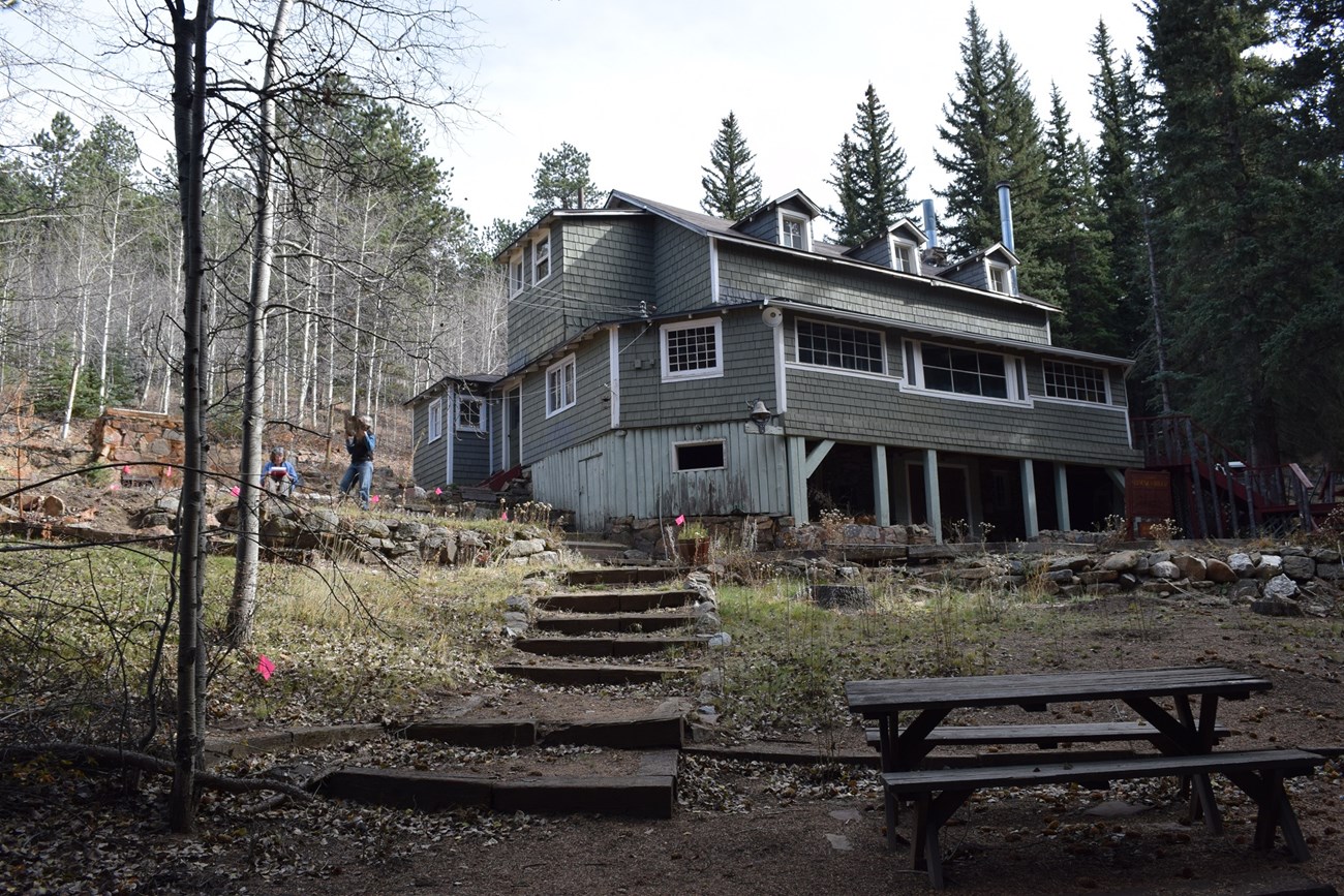 View of a two-and-a-half story building with basement from slope below while two individuals perform field survey at left