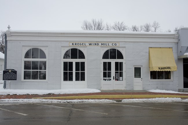A one-story building on a snowy street with two large doors and a sign that reads "Kregel Wind Mill Co."