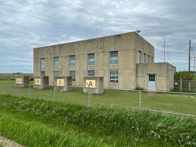 Reeve REA Power Generating Plant_08 Two-story power plant fronted by four air intake houses in a rural area. There are five large window openings at each level and three circled lightning bolts stamped into cornice.