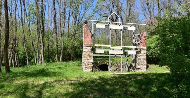 View of a metal frame outlining the form of a building now partially in ruins amid a green forested setting